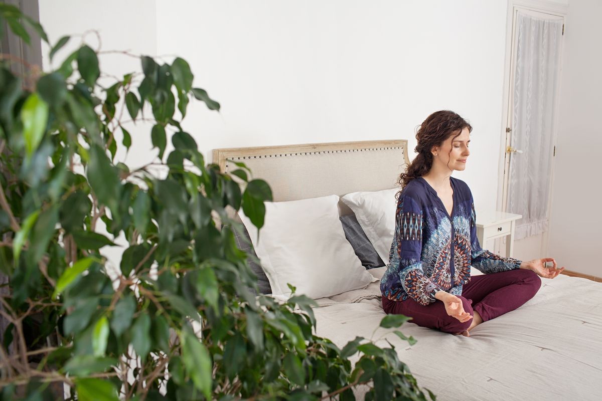 Side view of middle aged woman sitting on bed doing yoga during morning, home bedroom interior. Healthy female meditating, mind training, wellness mindful sport, indoors. Recreation lifestyle.
